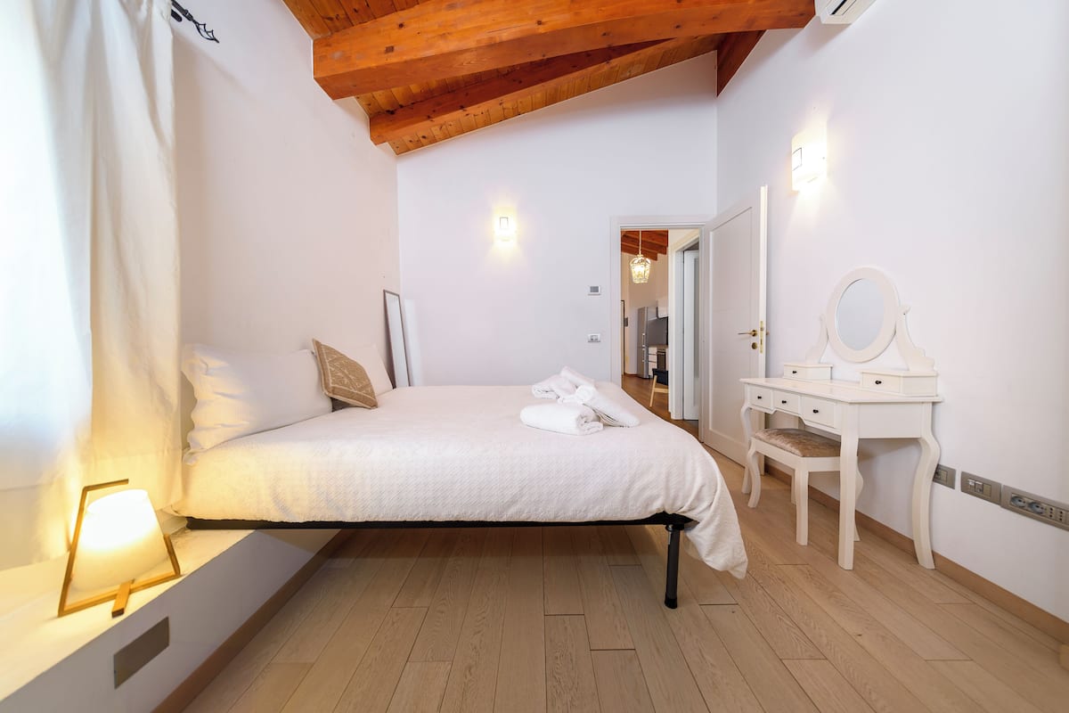 Quarto com cama, abajures, penteadeira com espelho e porta que dá para outro cômodo.