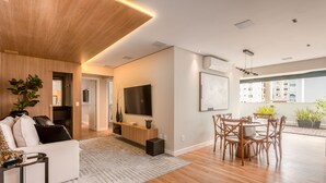 Una sala de estar moderna con techo de madera, un televisor de pantalla plana, un comedor con una mesa redonda y sillas, y un ventanal con vistas a edificios.