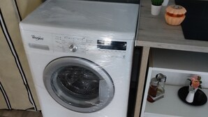 A compact, white Whirlpool washing machine in a modern kitchen with grey tiled backsplash and wooden flooring.