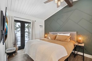 Ein Schlafzimmer mit einem Bett, einem Nachttisch mit Lampe, einem Fenster mit Vorhängen und einer zur Außenseite führenden Tür.