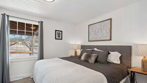 Une chambre à coucher avec un grand lit, une fenêtre avec des rideaux, une table de chevet avec une lampe et une œuvre d’art encadrée accrochée au mur.