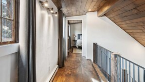 Een gang met houten vloer, een trap met metalen leuning en een zicht op een slaapkamer met een bed en nachtkastje.