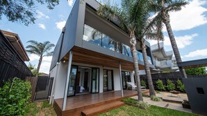 A modern two-story house with a wooden deck, glass doors, and a garden with palm trees. - Pet Friendly - Sandringham Sunshine Escape (Sandringham)