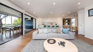 A modern living room with a grey sofa, a coffee table, and a dining area with wooden furniture. - Pet Friendly - Sandringham Sunshine Escape (Sandringham)