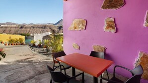 Una mesa y sillas frente a una pared morada con parches de piedra, con un paisaje montañoso al fondo.