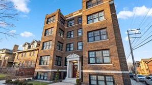 A multi-story brick building with a central entrance, large windows, and a parking area with cars. - Affordable 1-BR by DMC Detroit (Detroit)