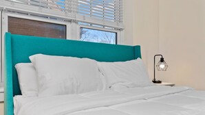 A bed with white bedding and a teal headboard, a nightstand with a black lamp, and a window with blinds. - Affordable 1-BR by DMC Detroit (Detroit)