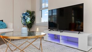 A modern living room with a glass-top coffee table, a flat-screen TV on a white entertainment unit, and a decorative vase with blue flowers. - Affordable 1-BR by DMC Detroit (Detroit)