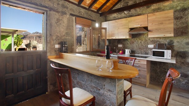 A rustic kitchen with a stone wall, wooden cabinets, a bar area, and a dining table with chairs. - Vacation Home 'Villa Paraiso Aldeano 1' with Mountain View, Shared Pool and Wi-Fi (La Aldea de San Nicolás)