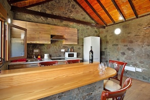 A rustic kitchen with wooden cabinets, a stone wall, a wooden dining table, and a white refrigerator.