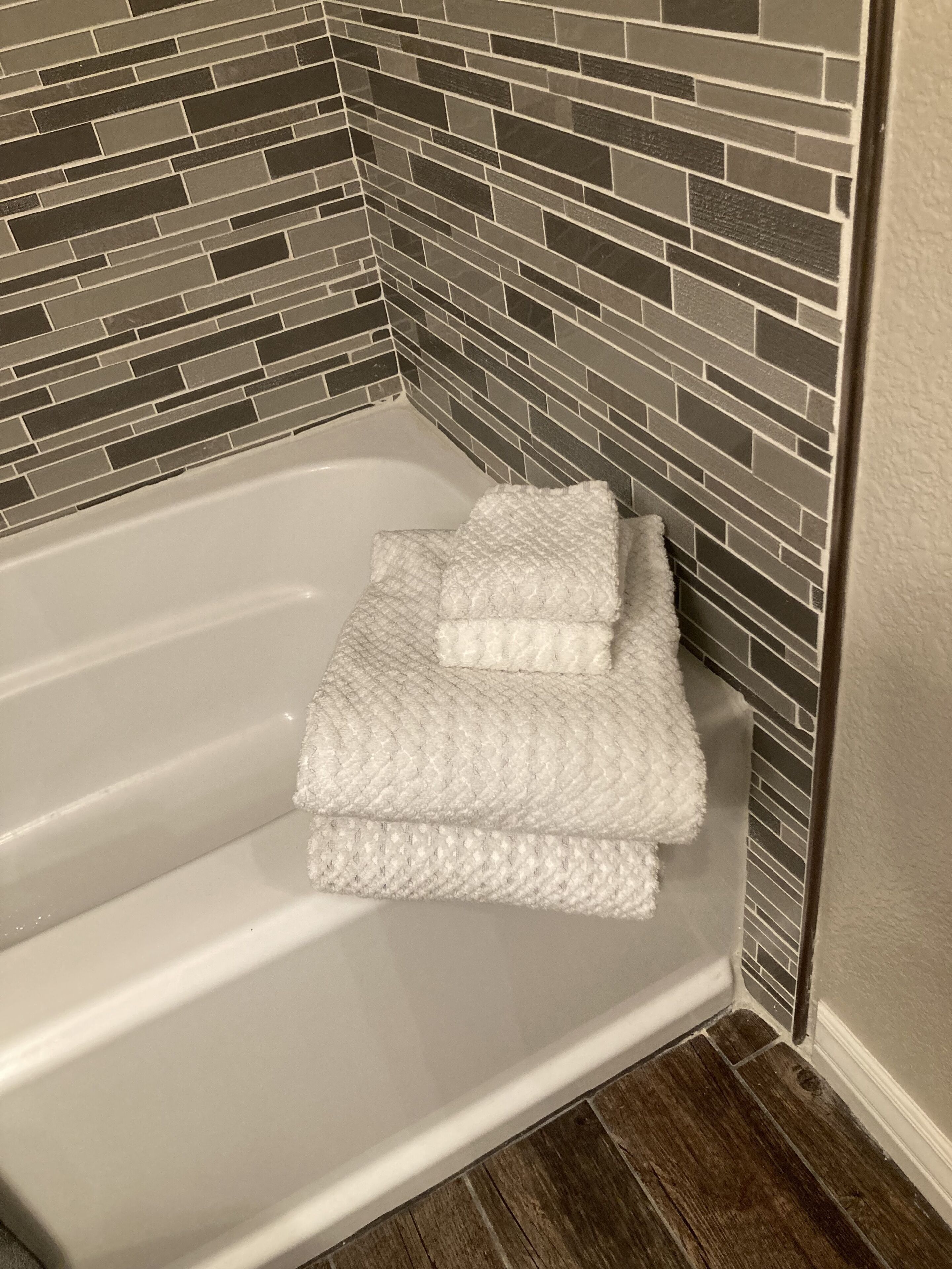 Ein Badezimmer-Eck mit einer weißen Badewanne, einem strukturierten weißen Handtuch und einer Wand mit Mosaikfliesen.
