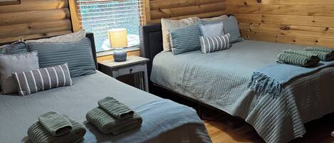 A cabin-style room with two beds, wooden walls, a window with blinds, and a small table with a lamp.