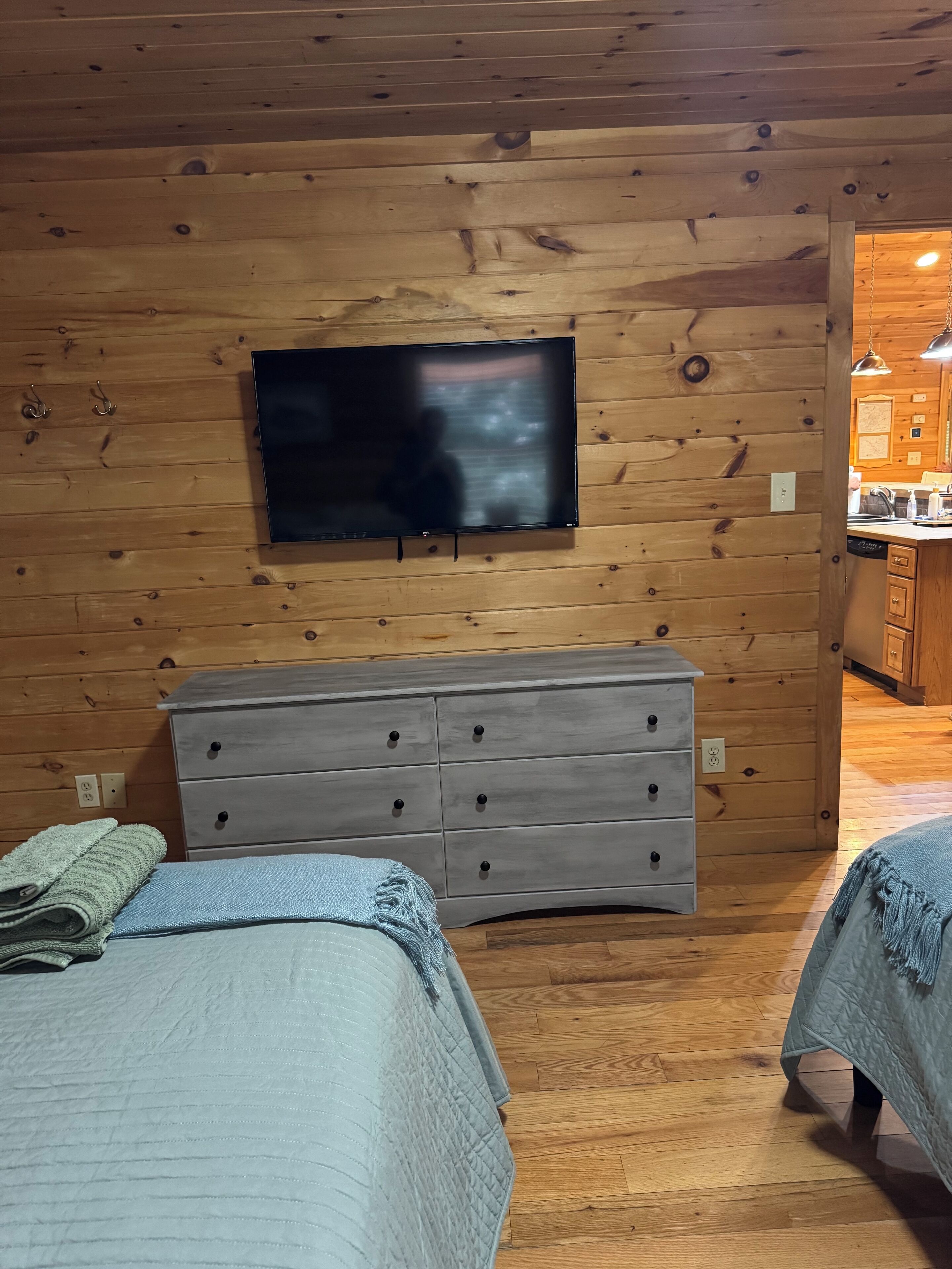 A cozy cabin room with wooden walls, a flat-screen TV mounted above a dresser, and two beds with blankets.