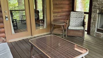 A wooden deck with a wicker table and chairs, a stone fireplace, and a glass door leading to an outdoor seating area.