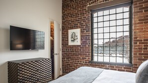 Een slaapkamer met een bakstenen muur, een raam met een ruitpatroon, een wandgemonteerde flatscreen-tv en een bed met een grijze deken.