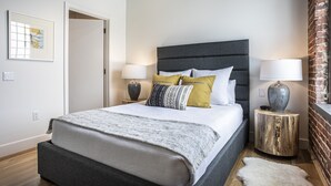 Een moderne slaapkamer met een groot bed, twee nachtkastjes met lampen, een lijst met een afbeelding aan de muur, en een bakstenen muur.