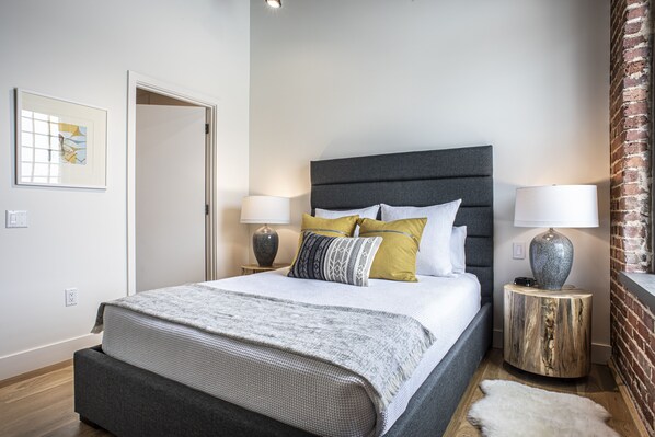 A modern bedroom with a large bed, two bedside tables with lamps, a framed picture on the wall, and a brick accent wall.