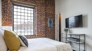 Een slaapkamer met een bakstenen muur, een groot raam, een bed met kussens, een televisie aan de muur en een metalen console tafel.