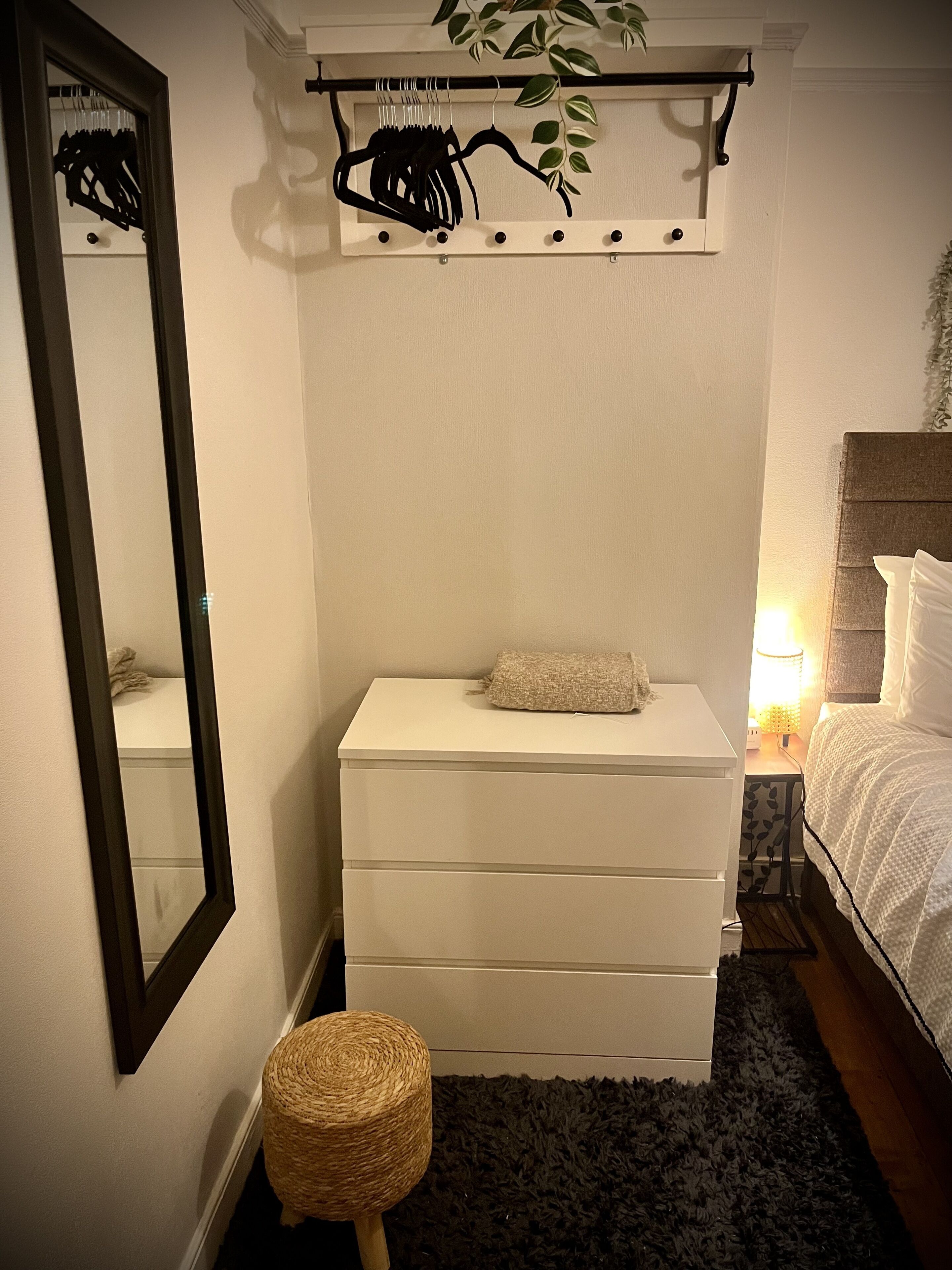Ein Schlafzimmer mit weißem Schminktisch, einem Spiegel, einem Nachttisch mit Lampe, einem Bett mit weißer Bettwäsche und einer hängenden Pflanze.