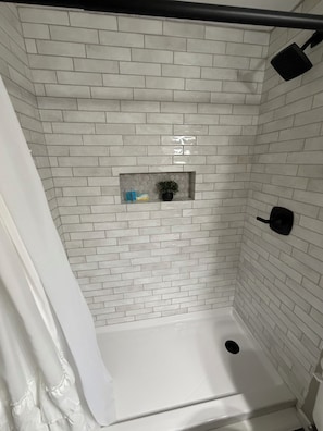 A shower with white tiled walls, a small shelf with toiletries, and a black shower head.