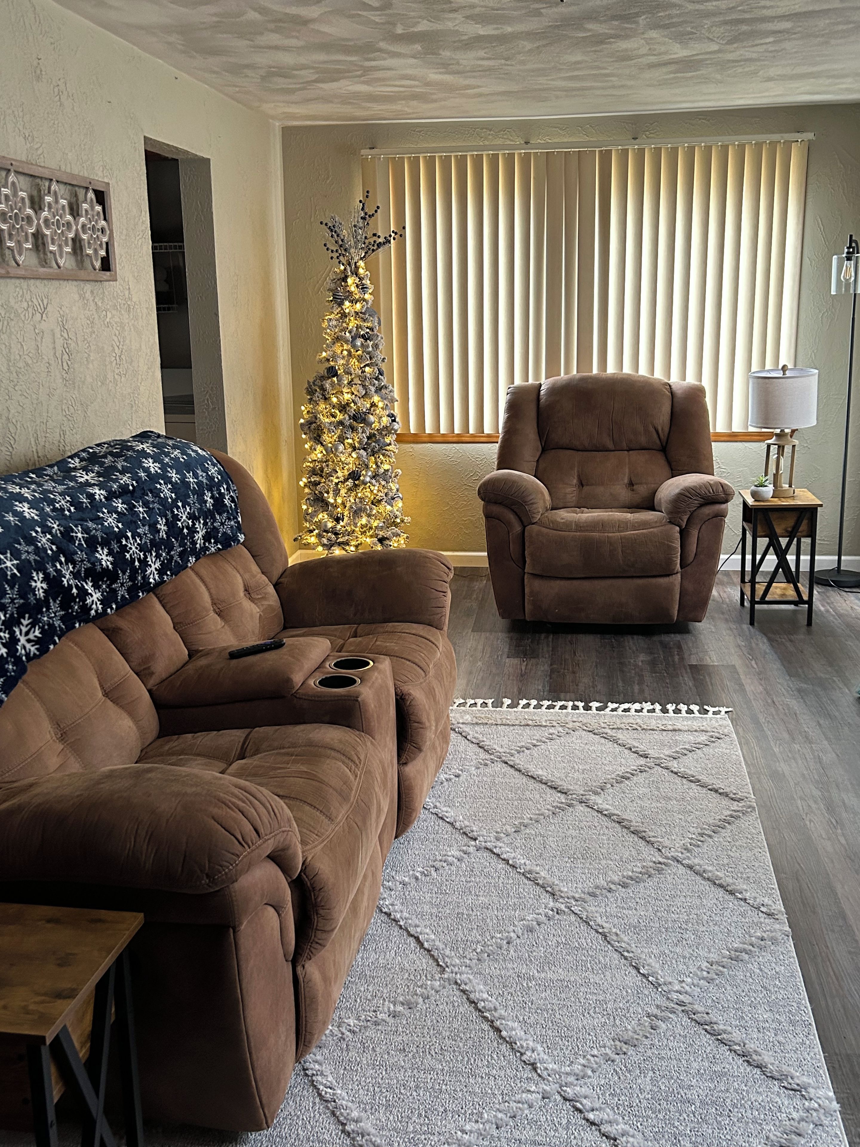 Una sala de estar con un sofá marrón, un sillón marrón, una mesita, un árbol decorativo y una lámpara de pie.