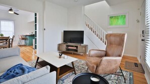 A modern living room with a flat-screen TV, a leather armchair, and a dining area with a table and chairs. - 3rd Ave Cottage - Close to Short North & Downtown (Columbus)
