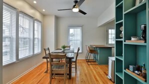 A dining area with a wooden table and chairs, a ceiling fan, and a kitchen area with a refrigerator and shelves. - 3rd Ave Cottage - Close to Short North & Downtown (Columbus)