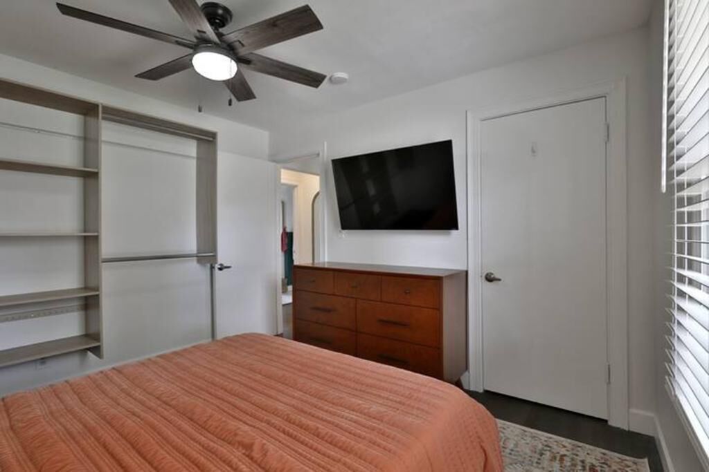 A bedroom with a bed, a ceiling fan, a TV mounted on the wall, a dresser, and a door.