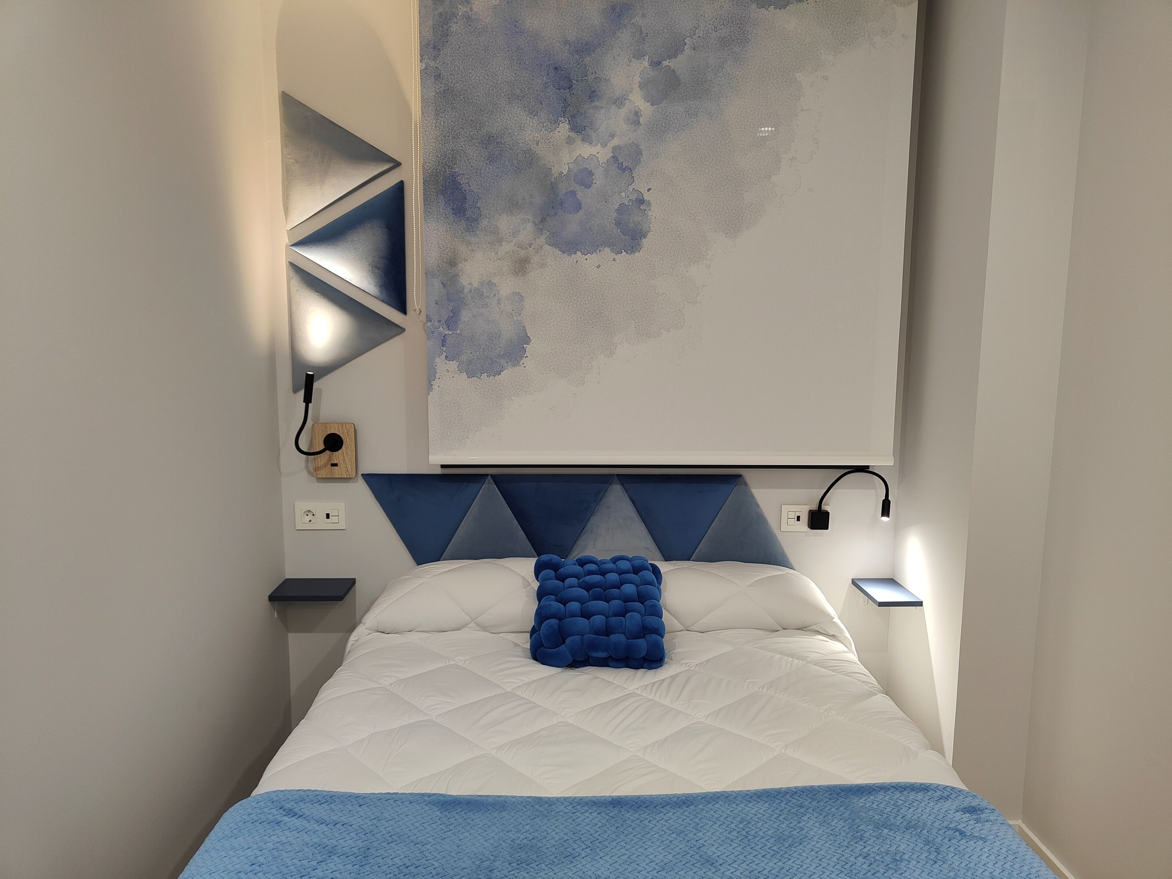 A bedroom with a bed, a blue headboard, a wall-mounted abstract painting, and a bedside table with a lamp.