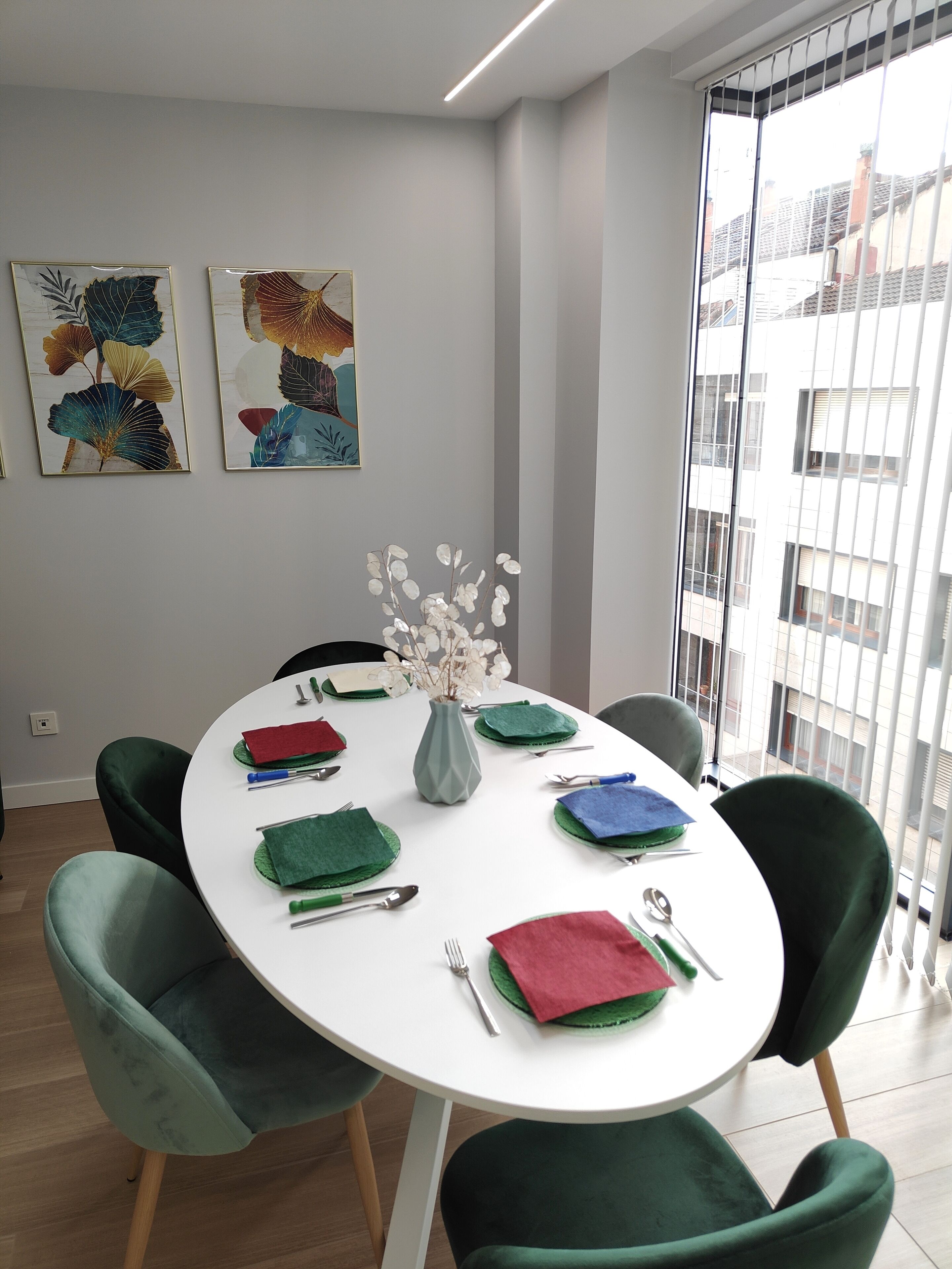 Un coin repas avec une table ronde dressée pour quatre personnes, des chaises à revêtement vert, un vase avec des fleurs blanches et deux œuvres d’art encadrées accrochées au mur.