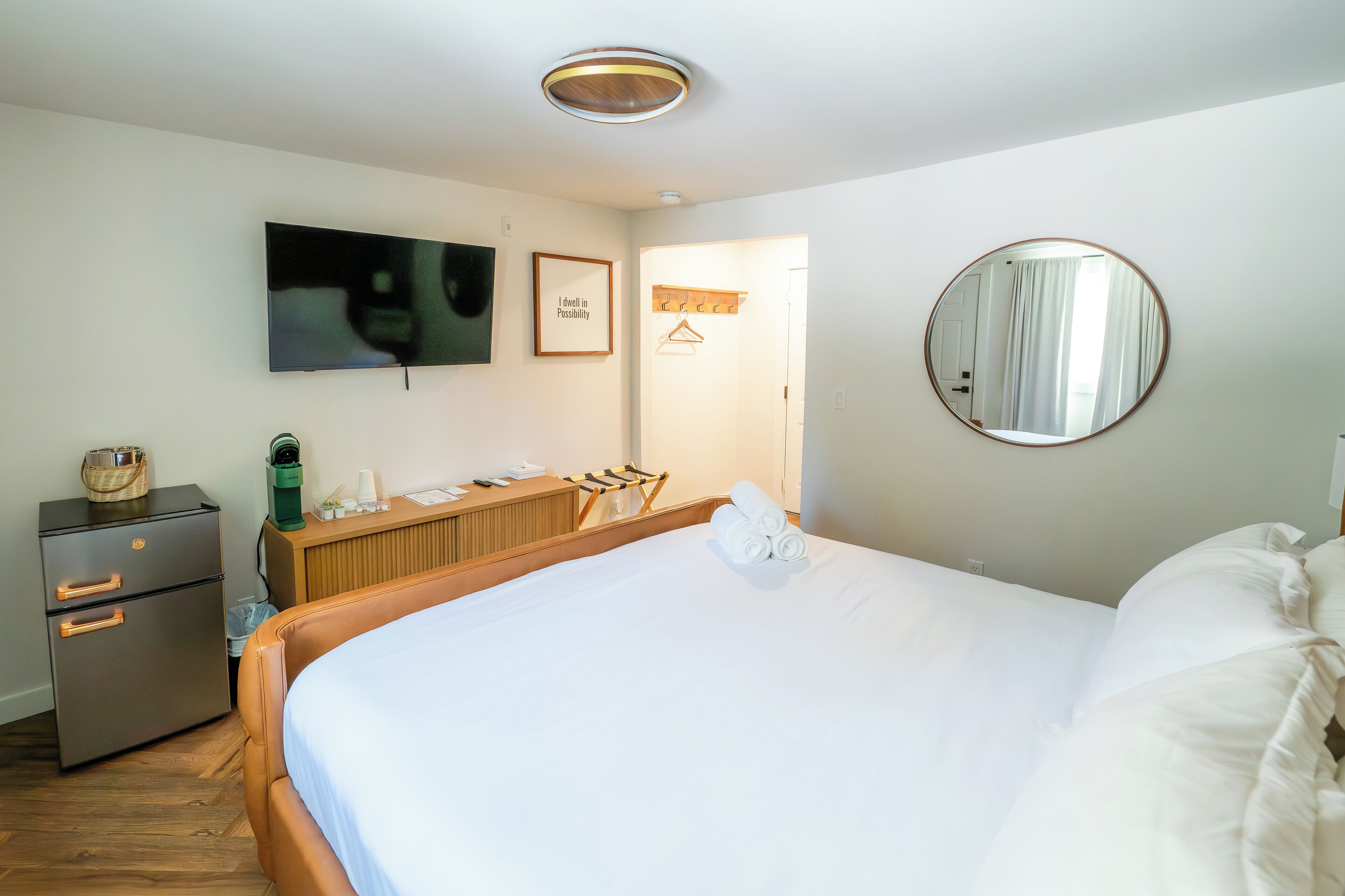 A hotel room with a large bed, a flat-screen TV mounted on the wall, a wooden dresser, a minibar, and a round mirror.