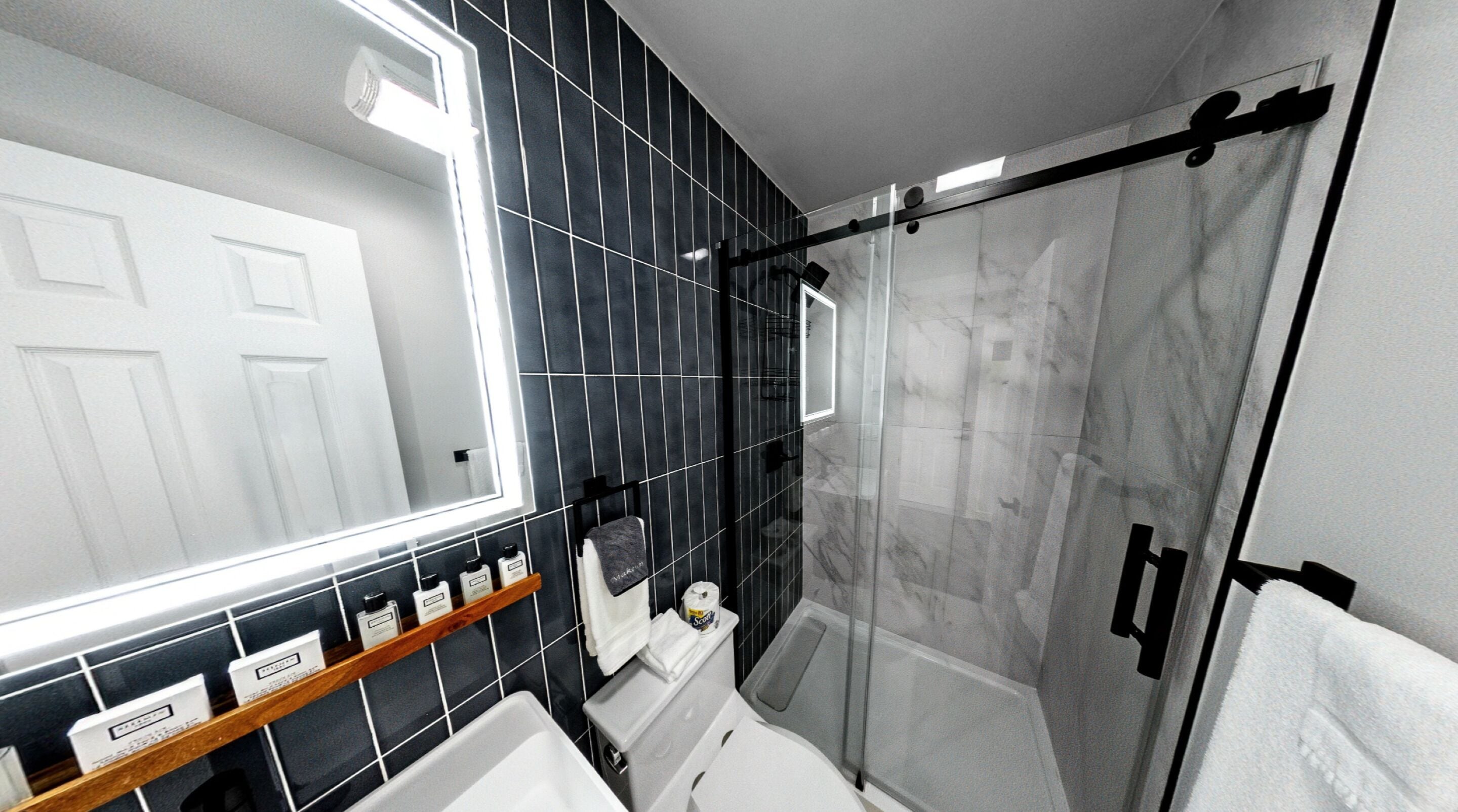 A modern bathroom with a large mirror, black tiled walls, a glass shower enclosure, and a white toilet.