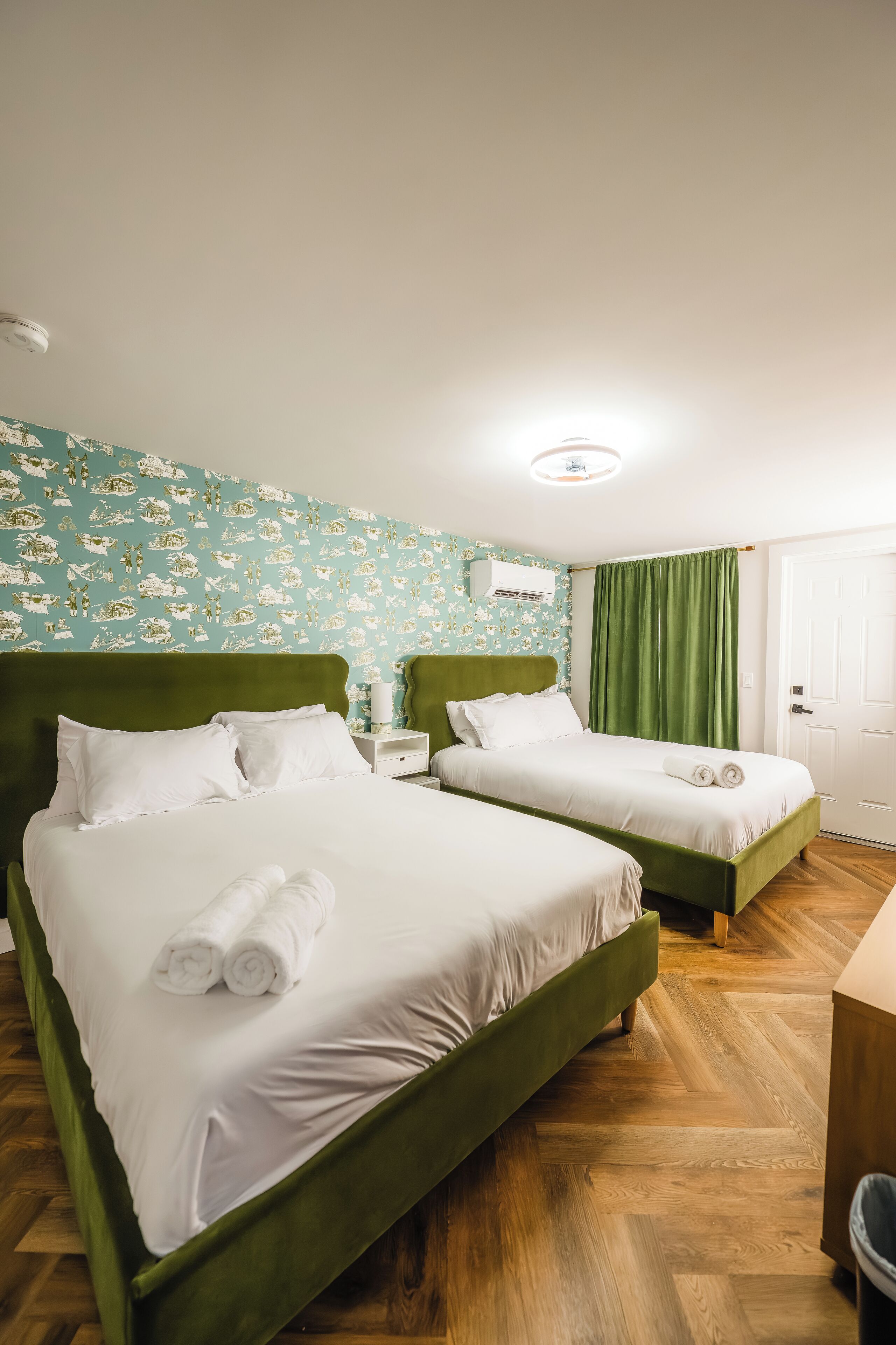 Habitación de hotel con dos camas, cabecera verde, ropa de cama blanca, pared con diseño y suelo de madera.