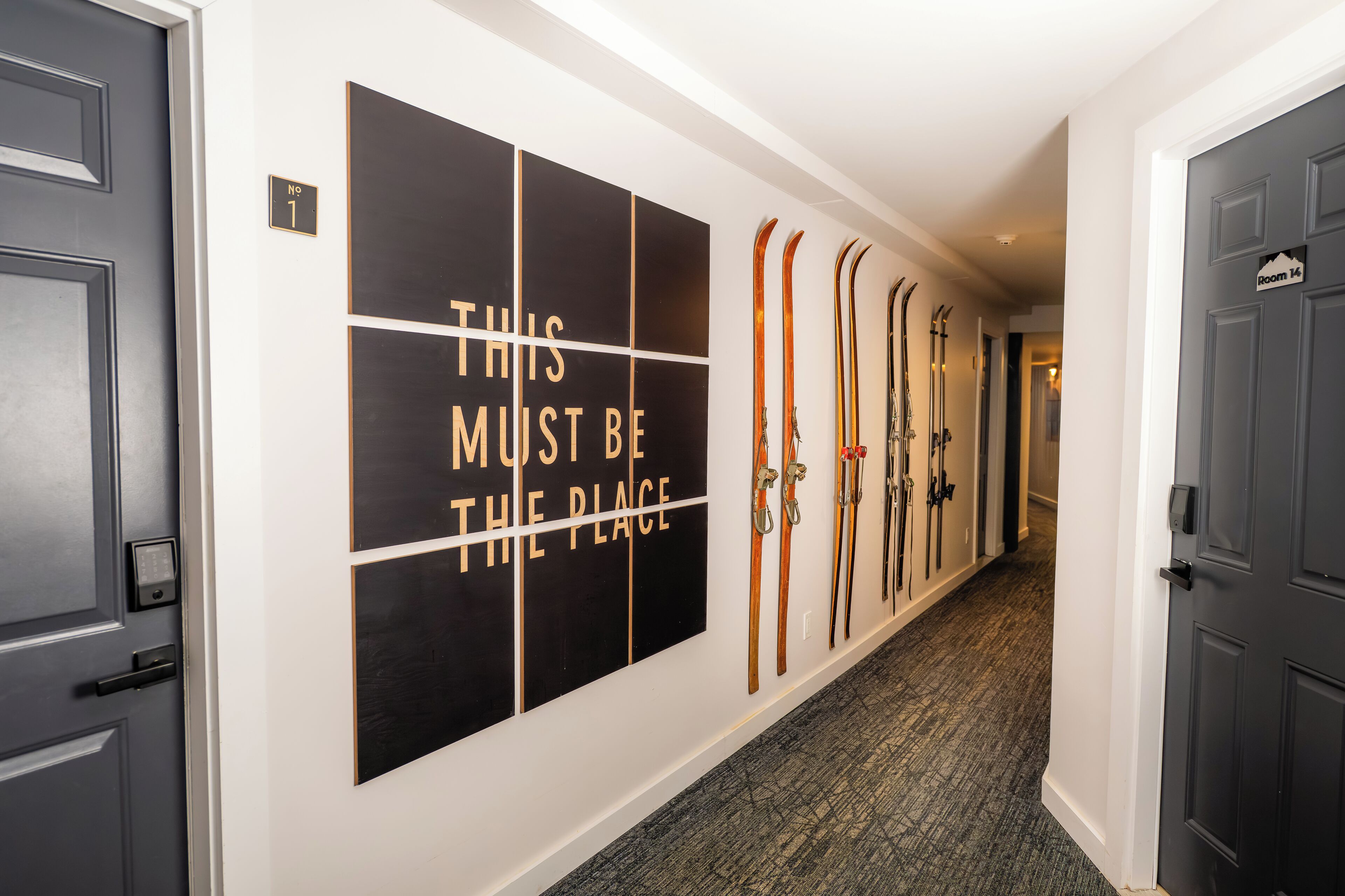 Un couloir avec une enseigne fixée au mur indiquant : « C’EST ICI » et une rangée de paires de skis accrochées au mur.