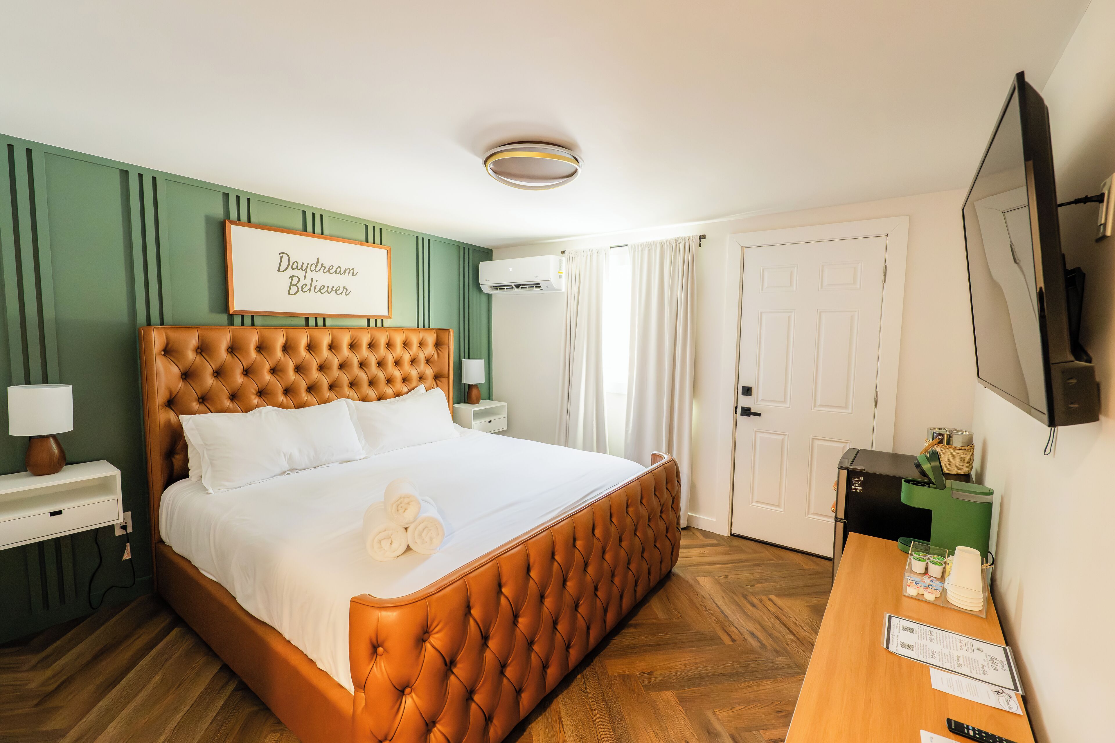 Ein Hotelzimmer mit einem bettenhohen Kopfende mit Wirbelung, einem Nachttisch mit Lampe, einem gerahmten Schild an der Wand und einem Schreibtisch mit Fernseher und einigen Gegenständen darauf.