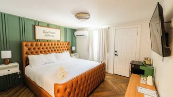 Ein Hotelzimmer mit einem bettenhohen Kopfende mit Wirbelung, einem Nachttisch mit Lampe, einem gerahmten Schild an der Wand und einem Schreibtisch mit Fernseher und einigen Gegenständen darauf.