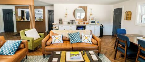 A modern living room with a brown leather sofa, a green armchair, and a wooden coffee table. There is a kitchen area with a microwave, toaster, and cabinets.