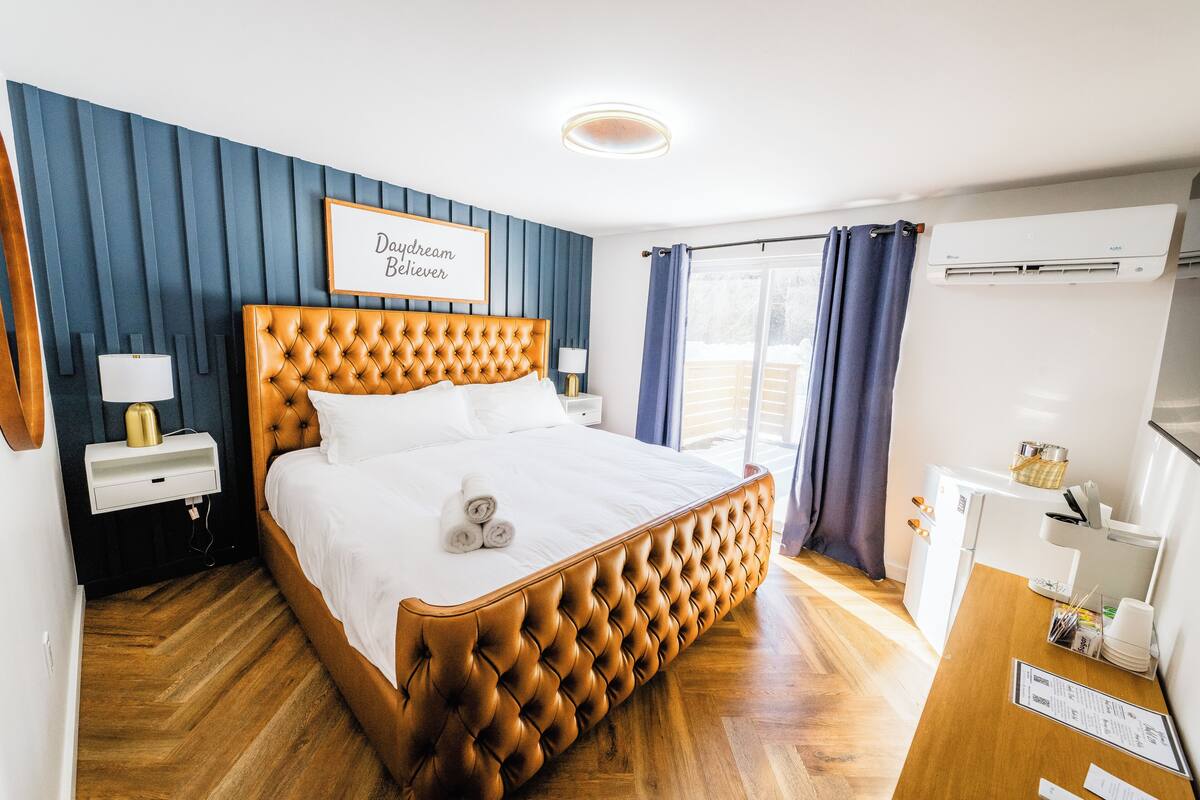 A neatly arranged bedroom with a tufted headboard, a bedside table with a lamp, and a window with curtains.