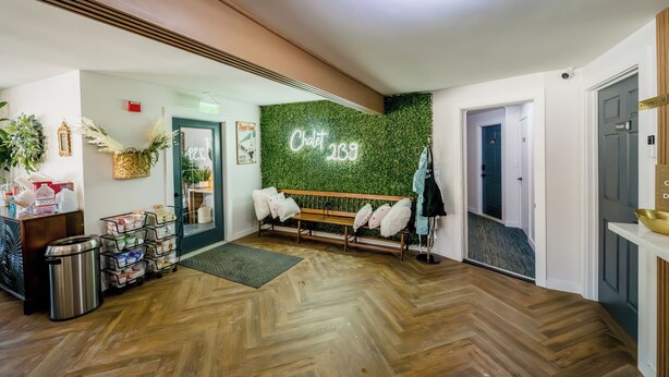 A modern hotel lobby with a wooden herringbone floor, a green wall with 'Chalet 289' signage, a bench with white cushions, and a reception desk with a trash bin and storage baskets. - Chalet 239 (West Dover)