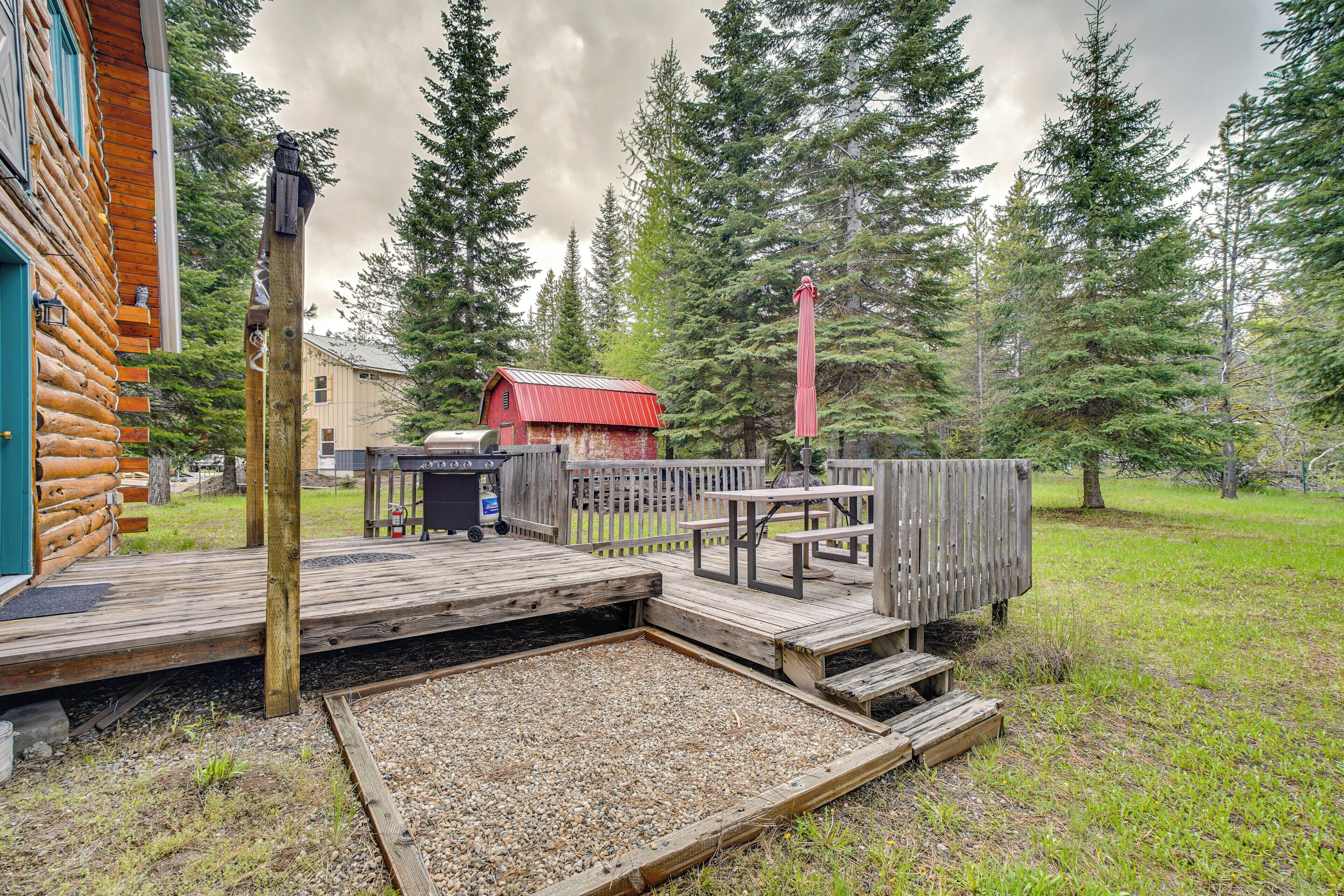 A wooden deck with picnic tables and a grill area, surrounded by a wooden fence and dense forest.