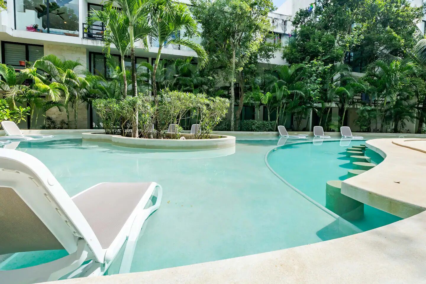 Una piscina con sillas de descanso, rodeada de plantas tropicales y un edificio al fondo.