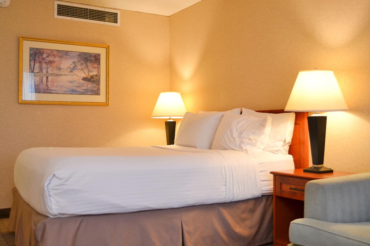 A hotel room with a bed, two bedside lamps, a nightstand, a chair, and a painting on the wall.