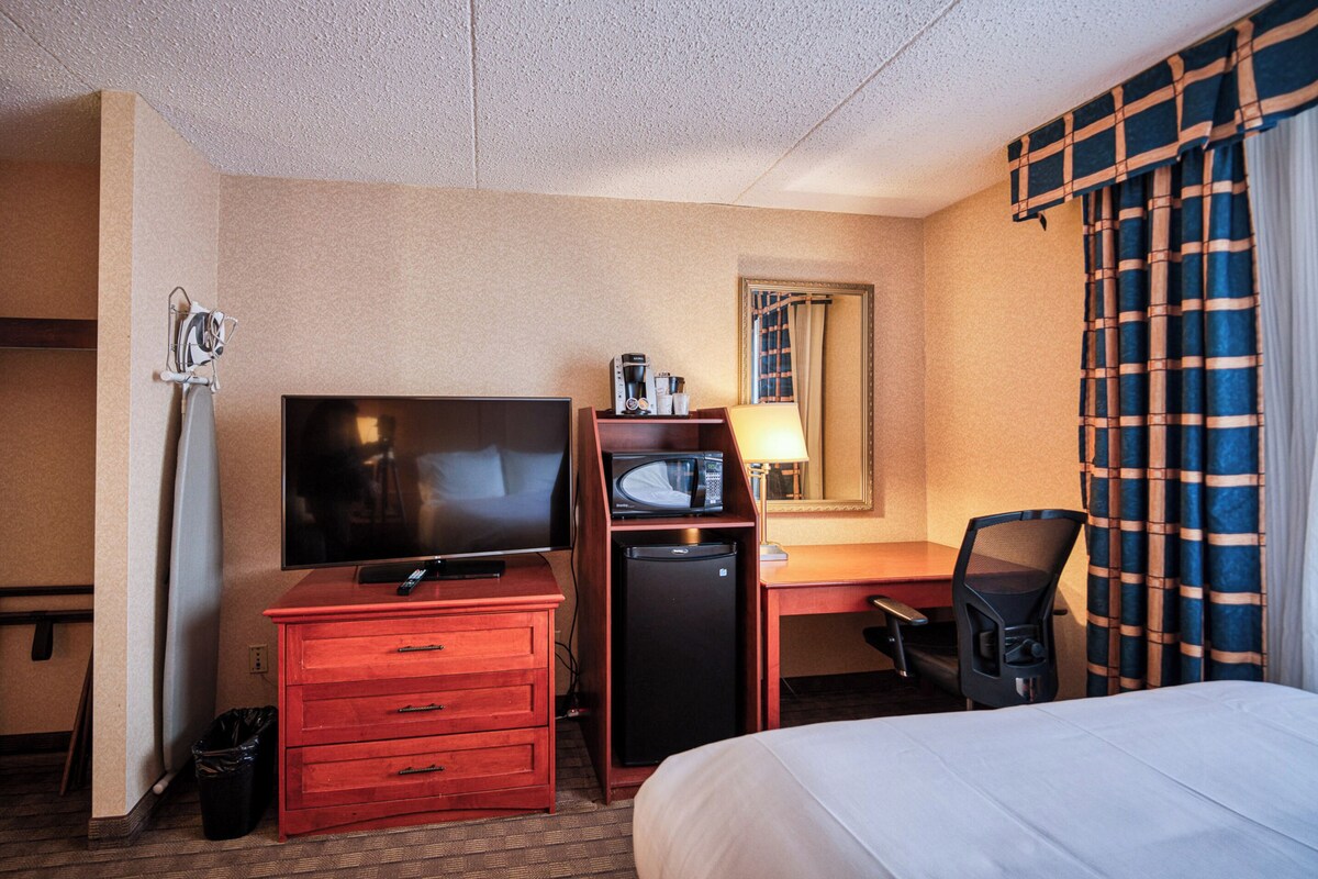 Habitación de hotel con cama, escritorio con computadora, televisión, microondas y bote de basura.