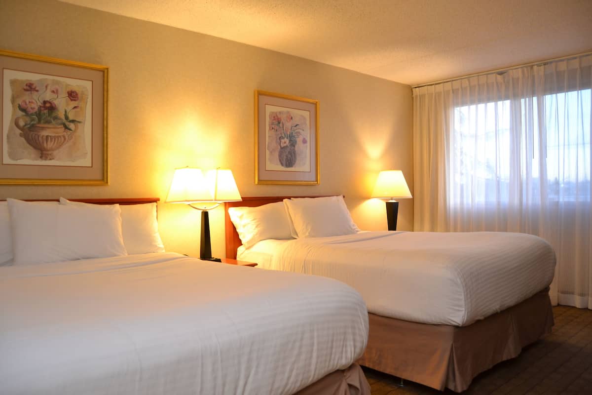 A hotel room with two beds, each with white linens, flanked by bedside tables with lamps. There are framed floral paintings on the wall above each bed.