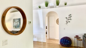 Ein modernes Wohnzimmer mit Holzboden, einem runden Spiegel, einem Gemälde, einem Hocker mit Pfauenmuster und einem an der Wand befestigten Pflanzenregal.