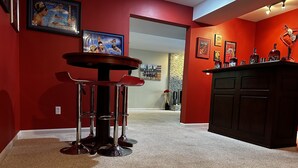 A room with a red wall, a round table with bar stools, a black cabinet with liquor bottles, and framed pictures on the wall.