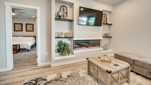 A modern living room with a fireplace, a flat-screen TV, and decorative shelves. - Hunt & Hike: Potholes State Park Home Hot Tub (Othello)