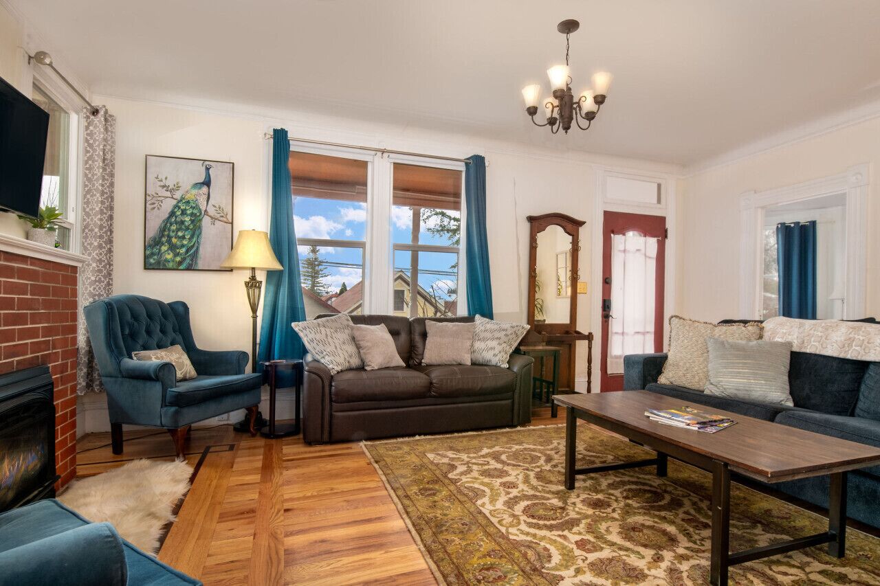 A living room with a fireplace, a blue armchair, a brown sofa, a wooden coffee table, and a large window with blue curtains.