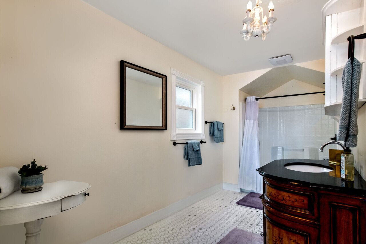 Une salle de bain avec un lavabo blanc, un grand miroir, un lustre et une fenêtre avec un porte-serviettes.