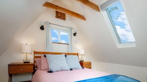Ein Schlafzimmer mit geneigter Decke, einem Oberlicht, einem Bett mit rosa und blauer Bettwäsche, zwei Nachttischen mit Lampen und einem hölzernen Kopfteil.